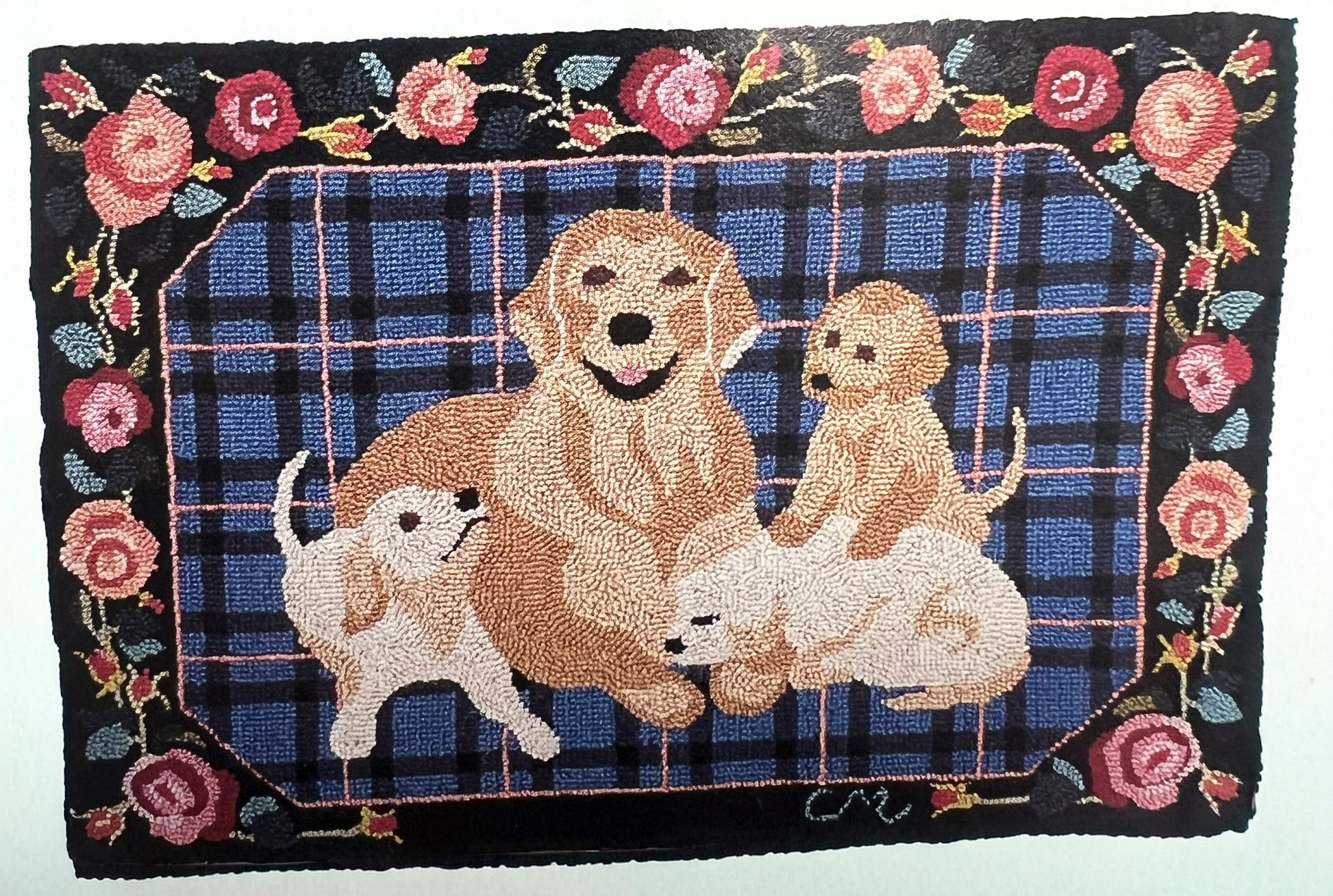 A rectangular hand-hooked rug featuring a family of golden retriever dogs in front of a blue plaid background, surrounded by a black border with pink roses and green leaves.