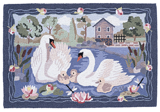 A hand-hooked rug featuring a family of white swans, two cygnets, reeds, water lilies, a small house, and a water wheel by a blue pond, bordered by a dark blue frame with pink flowers.