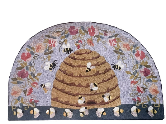 A semi-circular hand-hooked rug featuring a central tan beehive with bumblebees, surrounded by a floral border on a light lavender background and a dark gray base.
