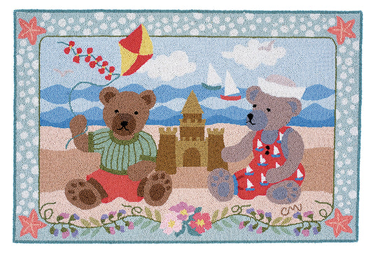 A hand-hooked rug featuring a whimsical beach scene with two teddy bears, a sandcastle, a red and yellow kite, mountains, and a floral border.
