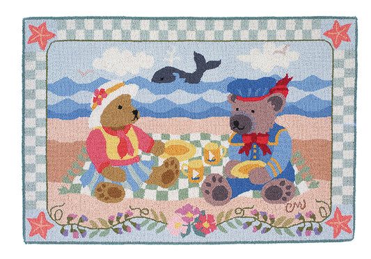 A hand-hooked rug featuring two teddy bears having a picnic on a beach. The left bear wears a pink and yellow outfit, and the right bear wears a blue outfit with a red bandana.  The ocean in the background with a whale.