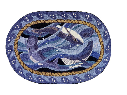 An oval hand-hooked rug featuring a blue whales swimming in the center, bordered by a rope motif and white and blue waves  on a light blue/gray background.